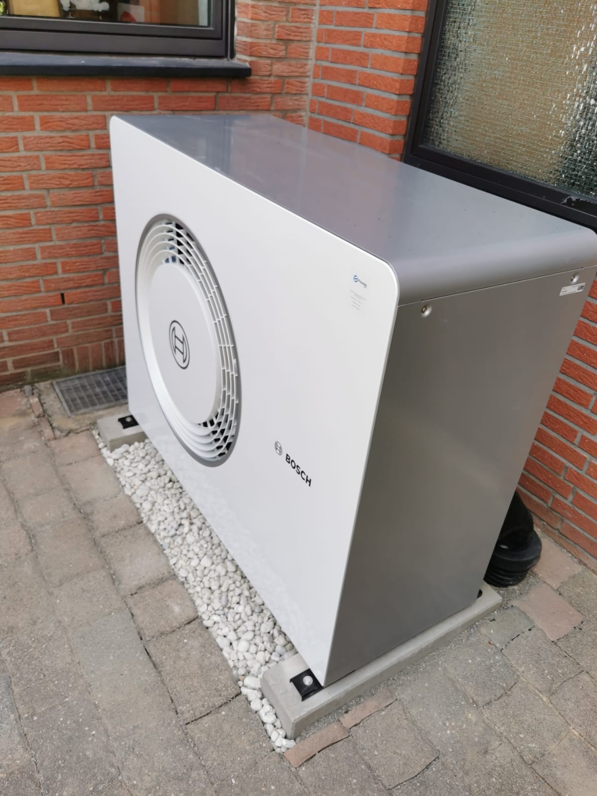 Bosch-Wärmepumpe vor roter Ziegelfassade auf gepflastertem Hof mit Kiesbett und Glasfenster im Hintergrund.